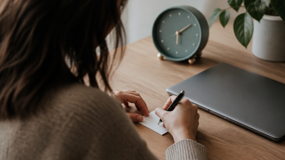 Worker makes a brief note on a small card beside a closed laptop and clock at a home workspace.